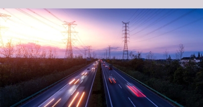Autopista al anochecer, cuatro carriles, ida y vuelta. Vehículos con los faros encendidos. Fondo de torretas eléctricas. Fuente: WTW